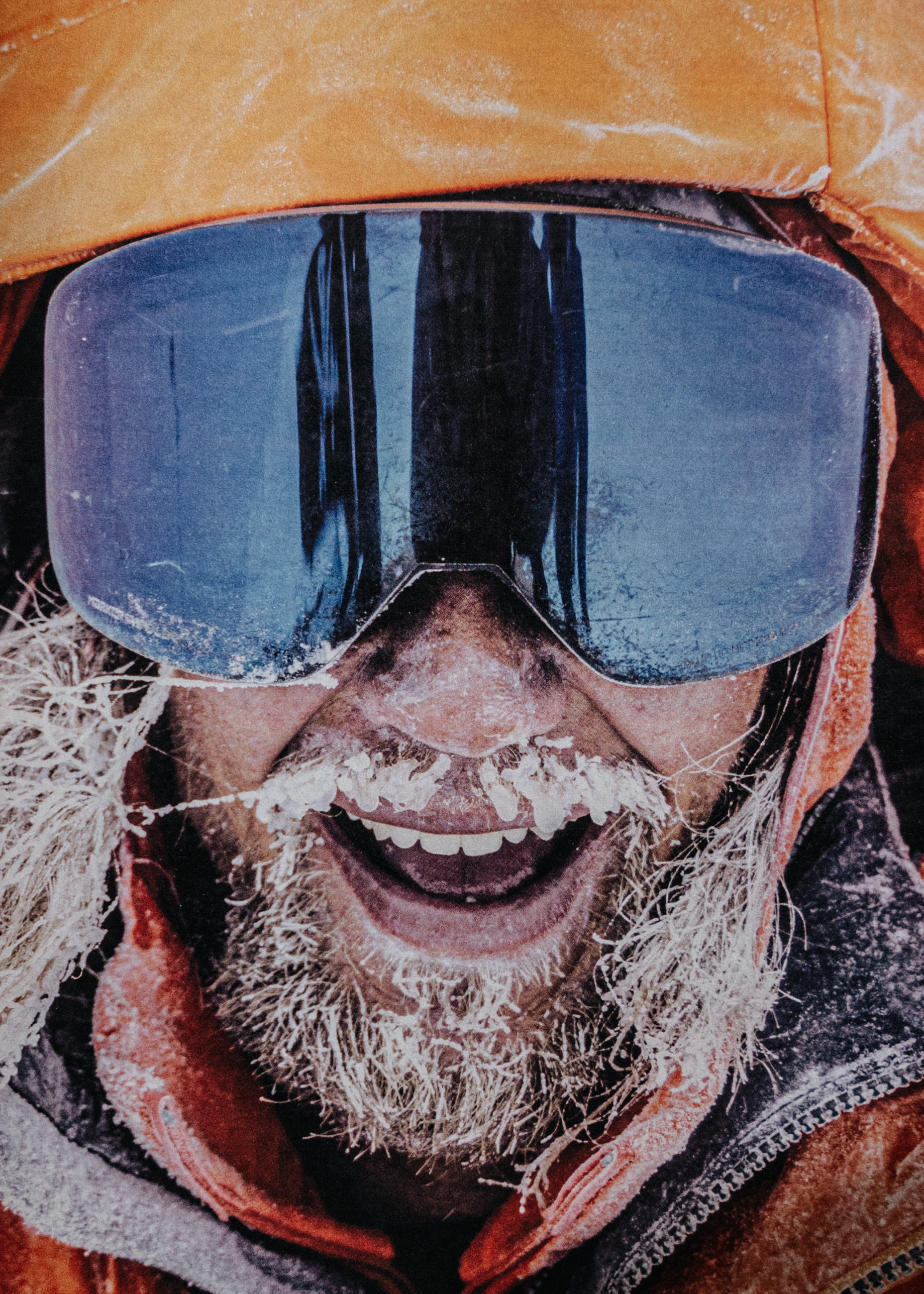 Smiling person with frost-covered beard wearing large ski goggles and an orange hooded jacket.