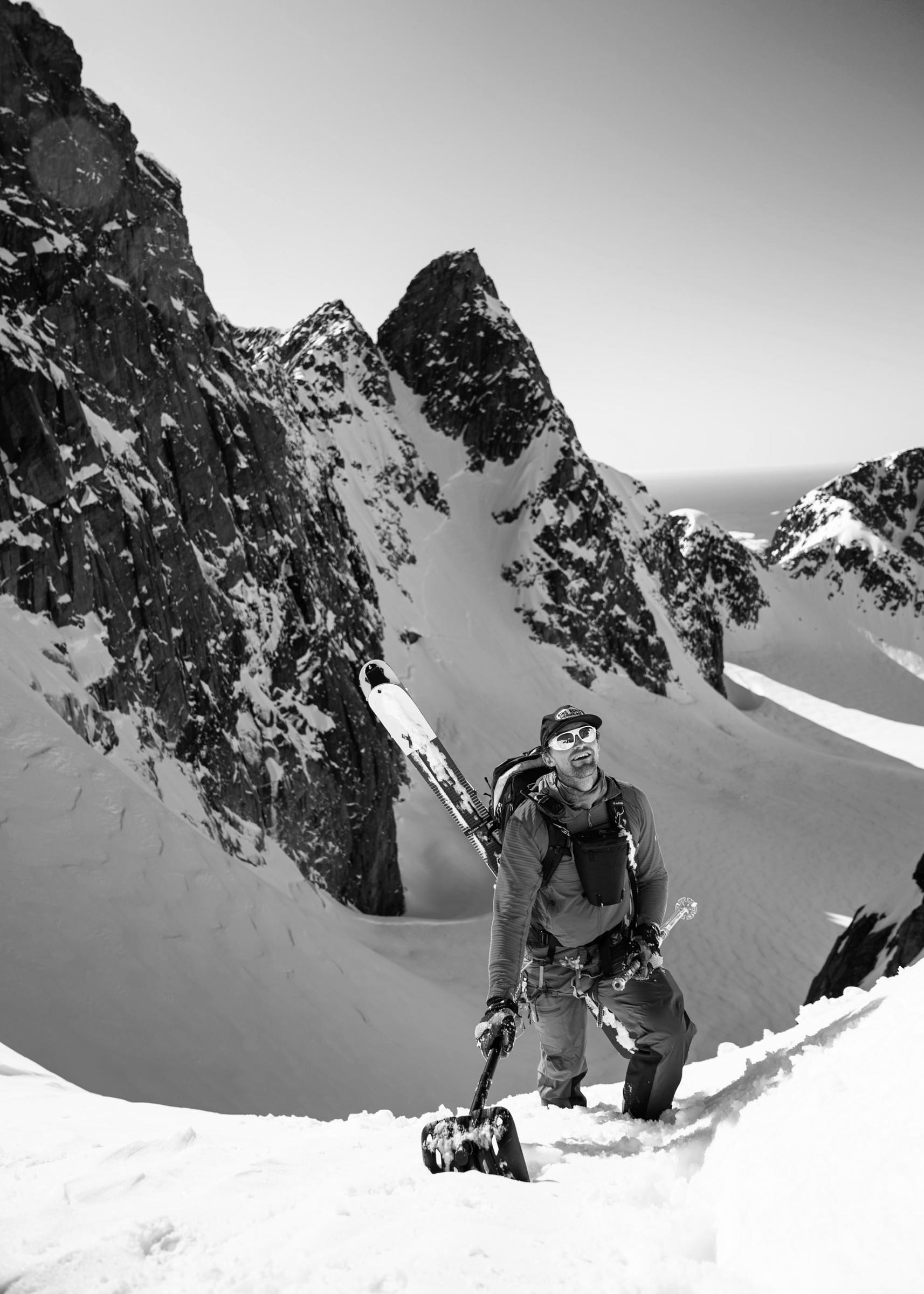 Man with skis and backpack climbing snowy mountain slope with shovel amid rocky peaks.