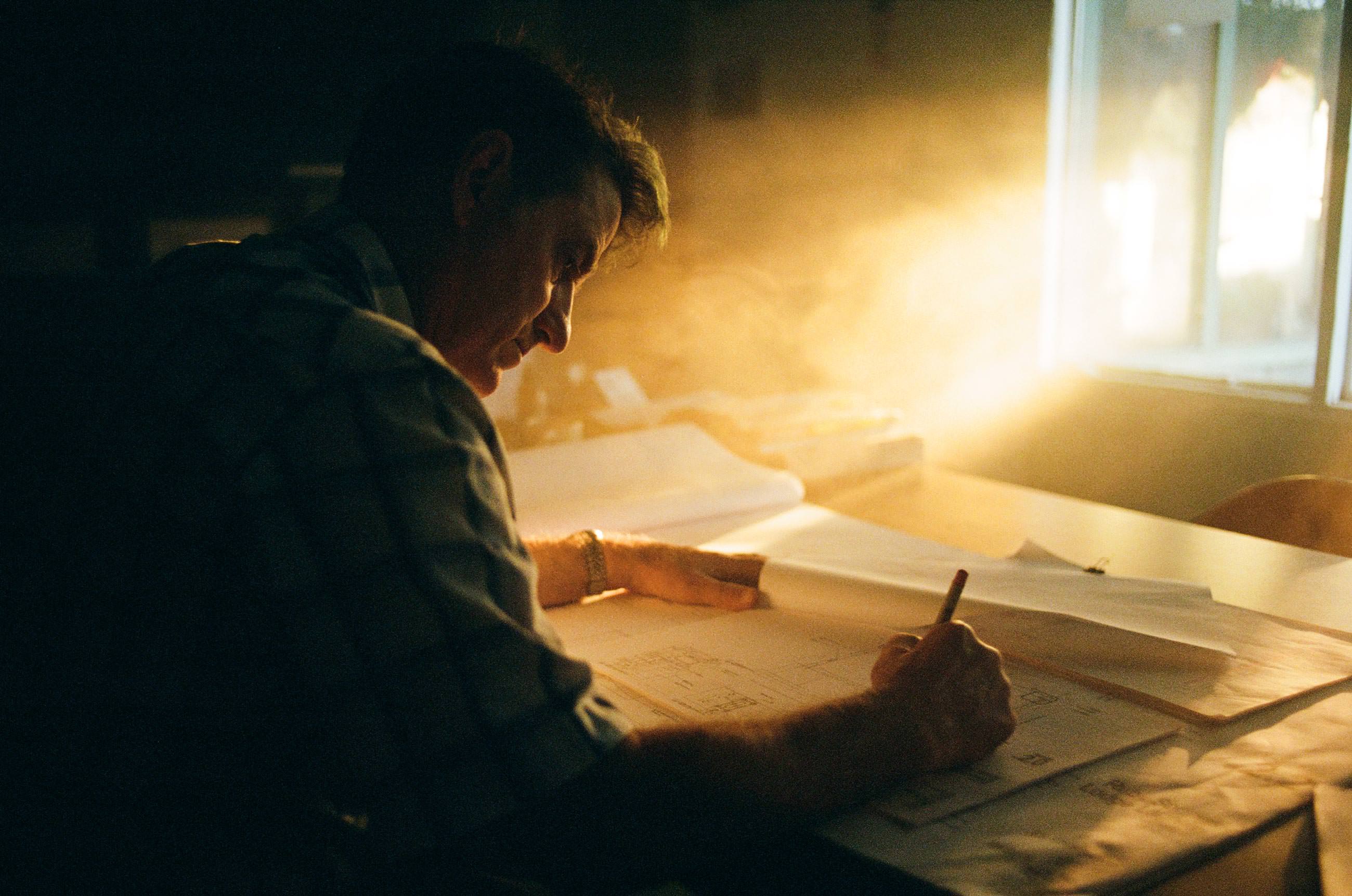 Man drawing plans on large paper at a desk with sunlight streaming through a window.