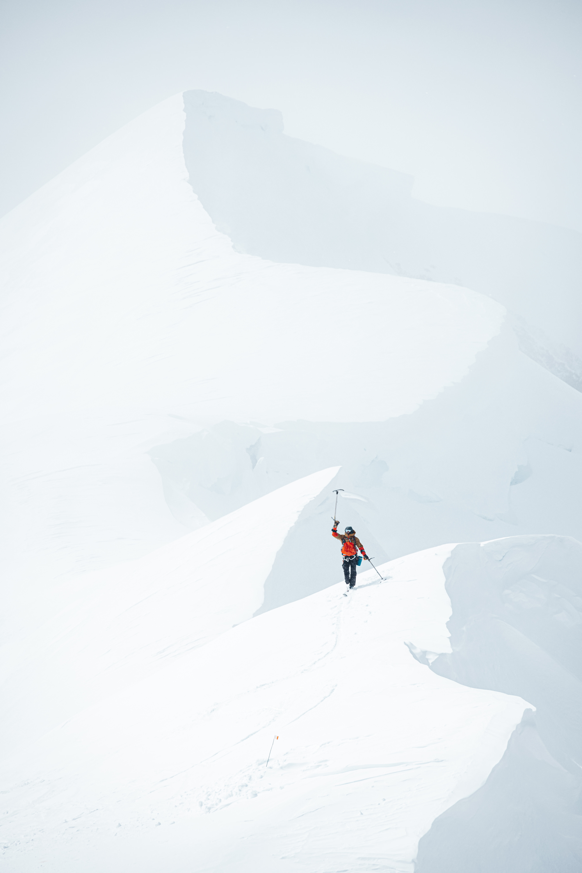 Skier in orange jacket raising an ice axe while standing on a snow-covered mountain ridge.