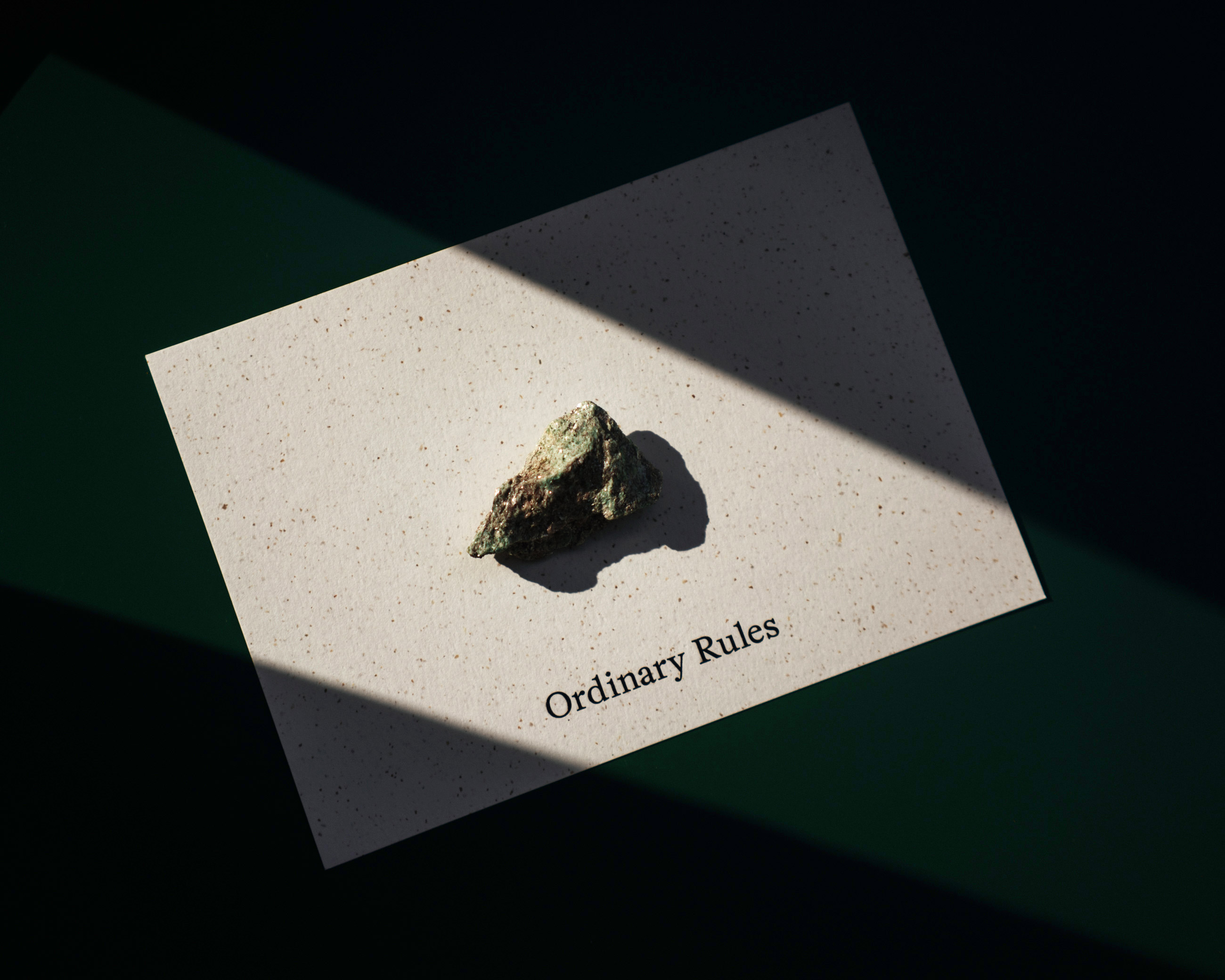 A textured stone on a speckled card with the words 'Ordinary Rules' under angled light beams.