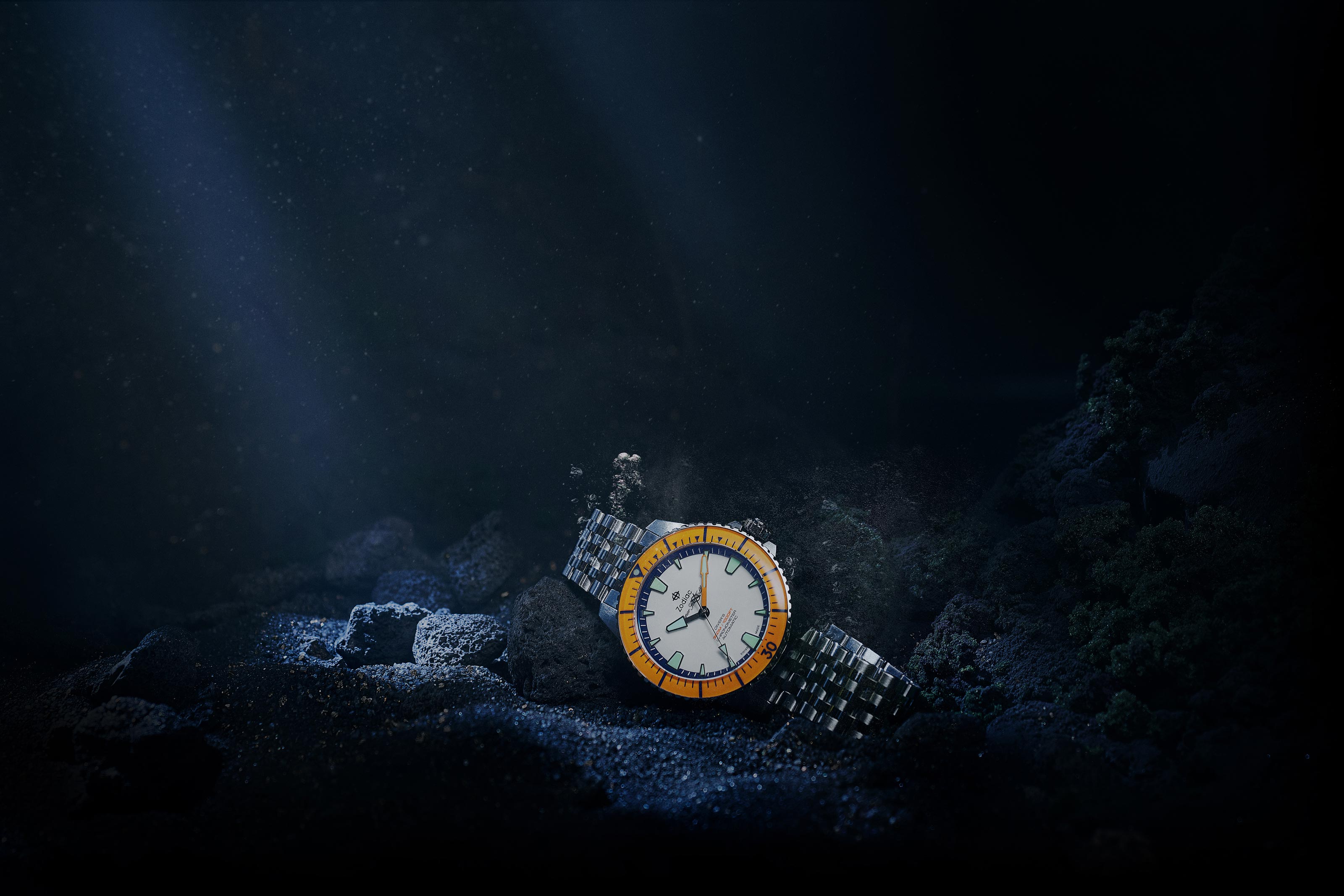 Silver wristwatch with an orange bezel resting on dark rocky seabed with bubbles rising.
