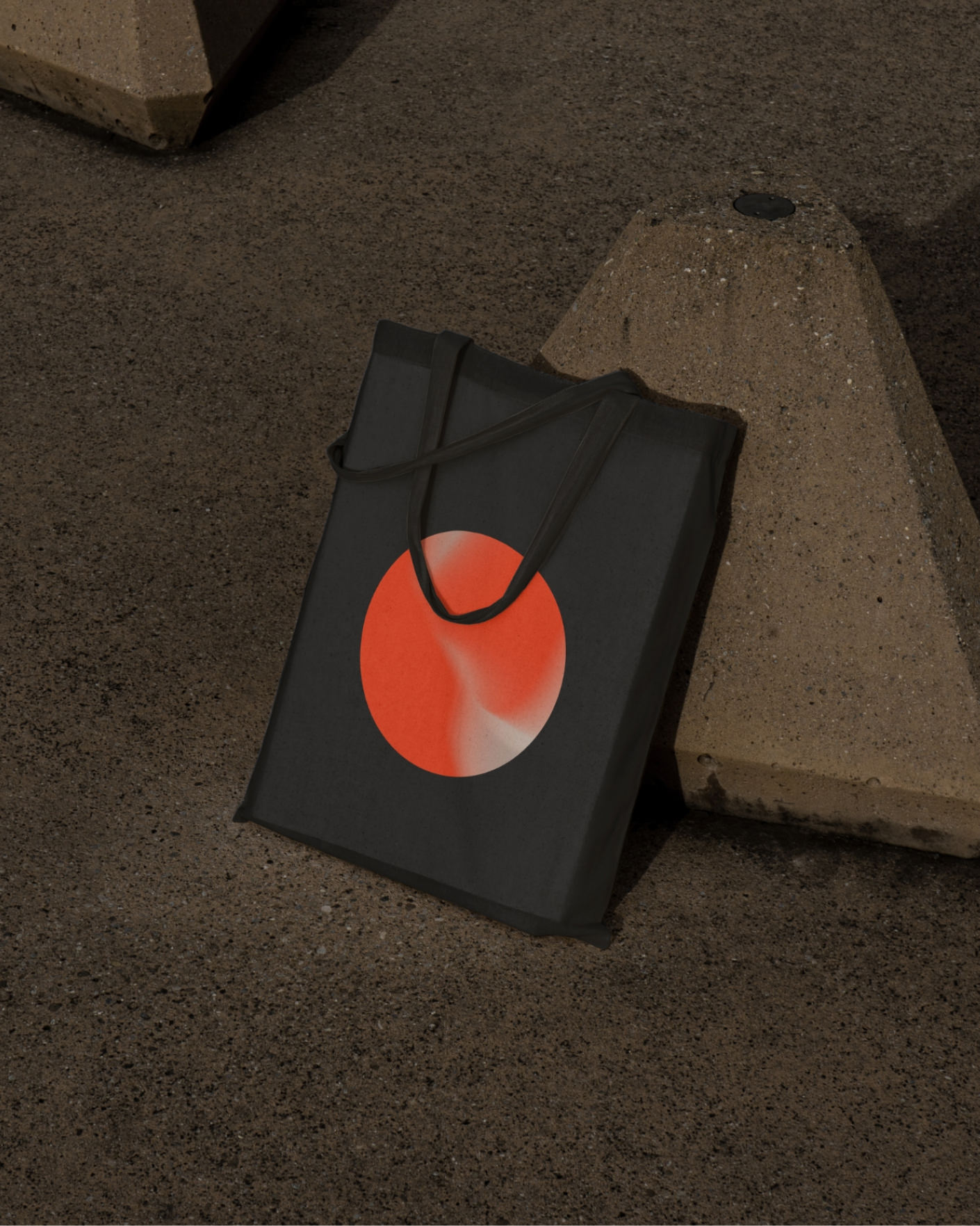 Black tote bag with red and white circular design resting against a concrete block on rough ground.