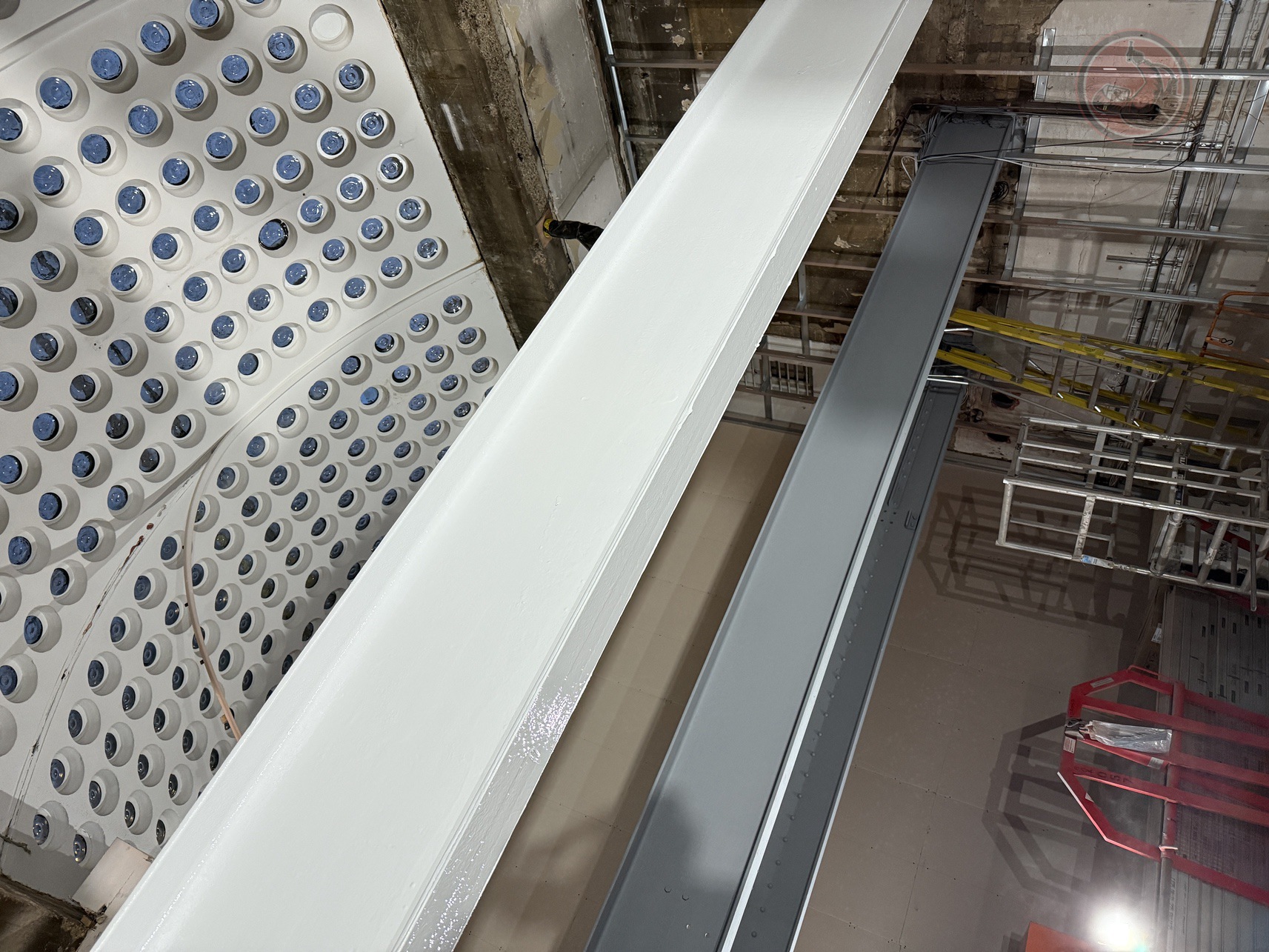 Interior construction site featuring painted steel beams, scaffolding, ladders, and a ceiling with circular recessed lights.