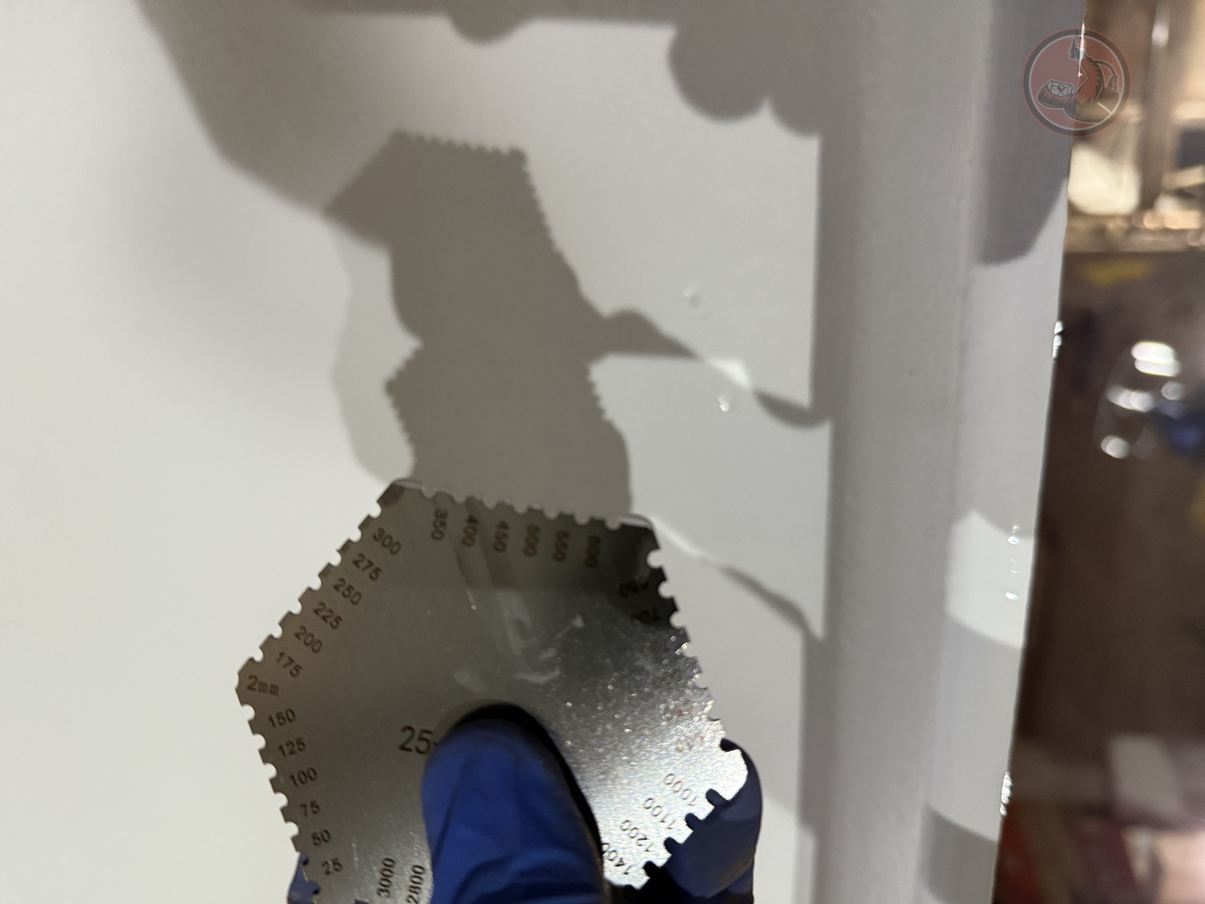 Hand wearing a blue glove holding a metal wet film thickness gauge with various notch sizes against a white surface.