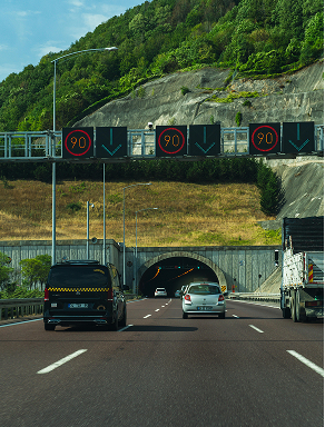 Highway tunnel entrance with cars and trucks, speed limit signs showing 90 km/h and green downward arrows above lanes.