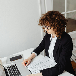 Woman with curly hair and glasses working on a laptop with documents on a white desk.