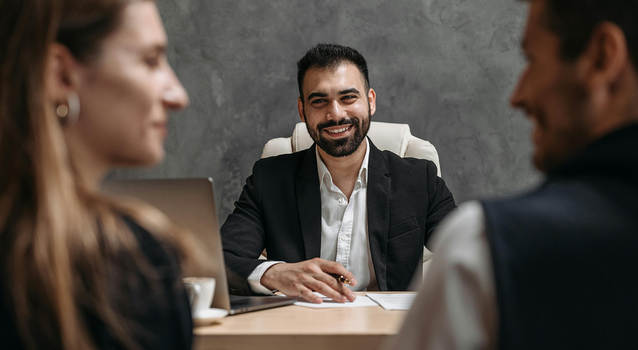Smiling male professional in a black blazer sitting at desk with laptop and documents, talking to two clients.