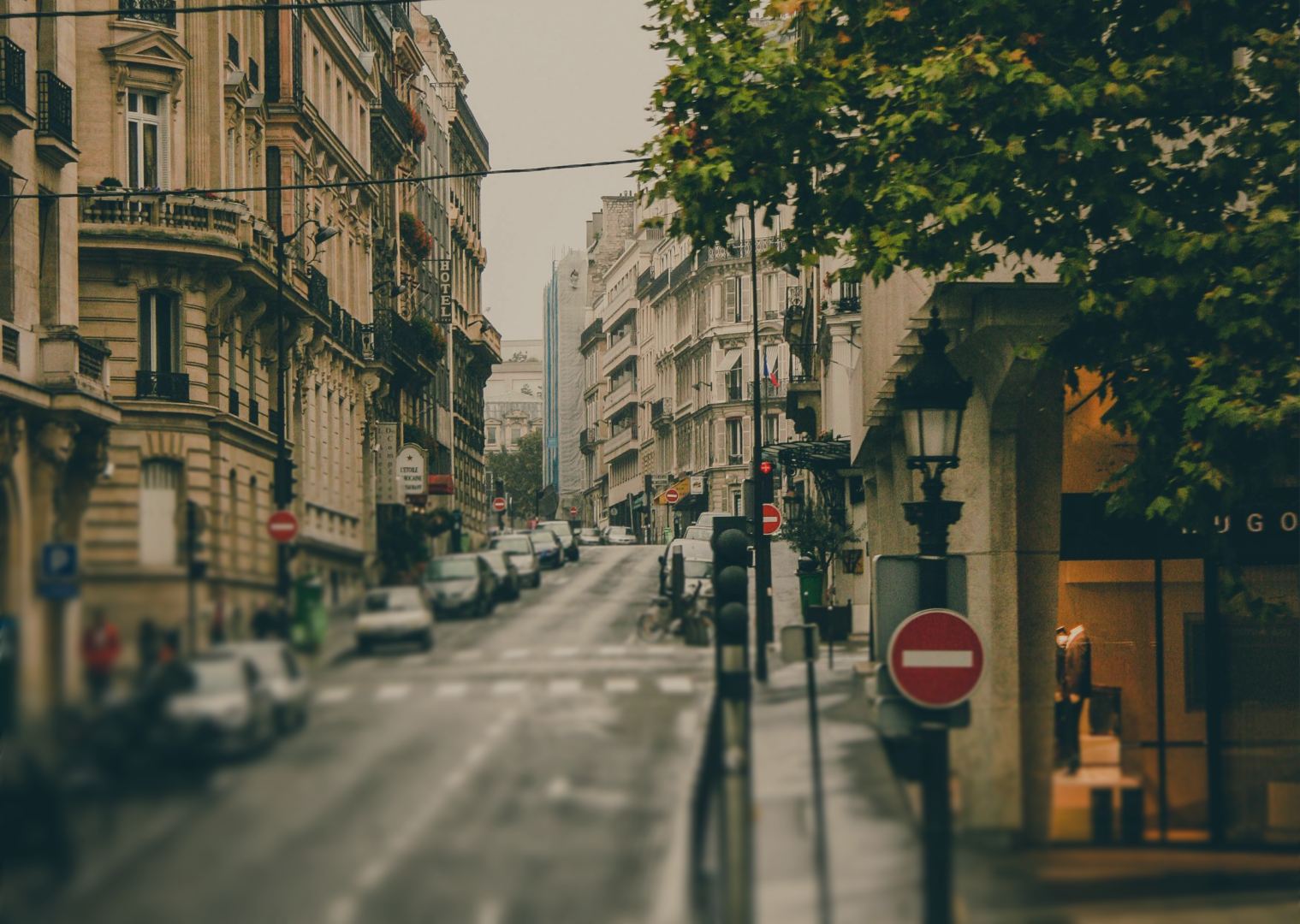 Street view of a European city with classic buildings, parked cars, traffic signs, and green trees.