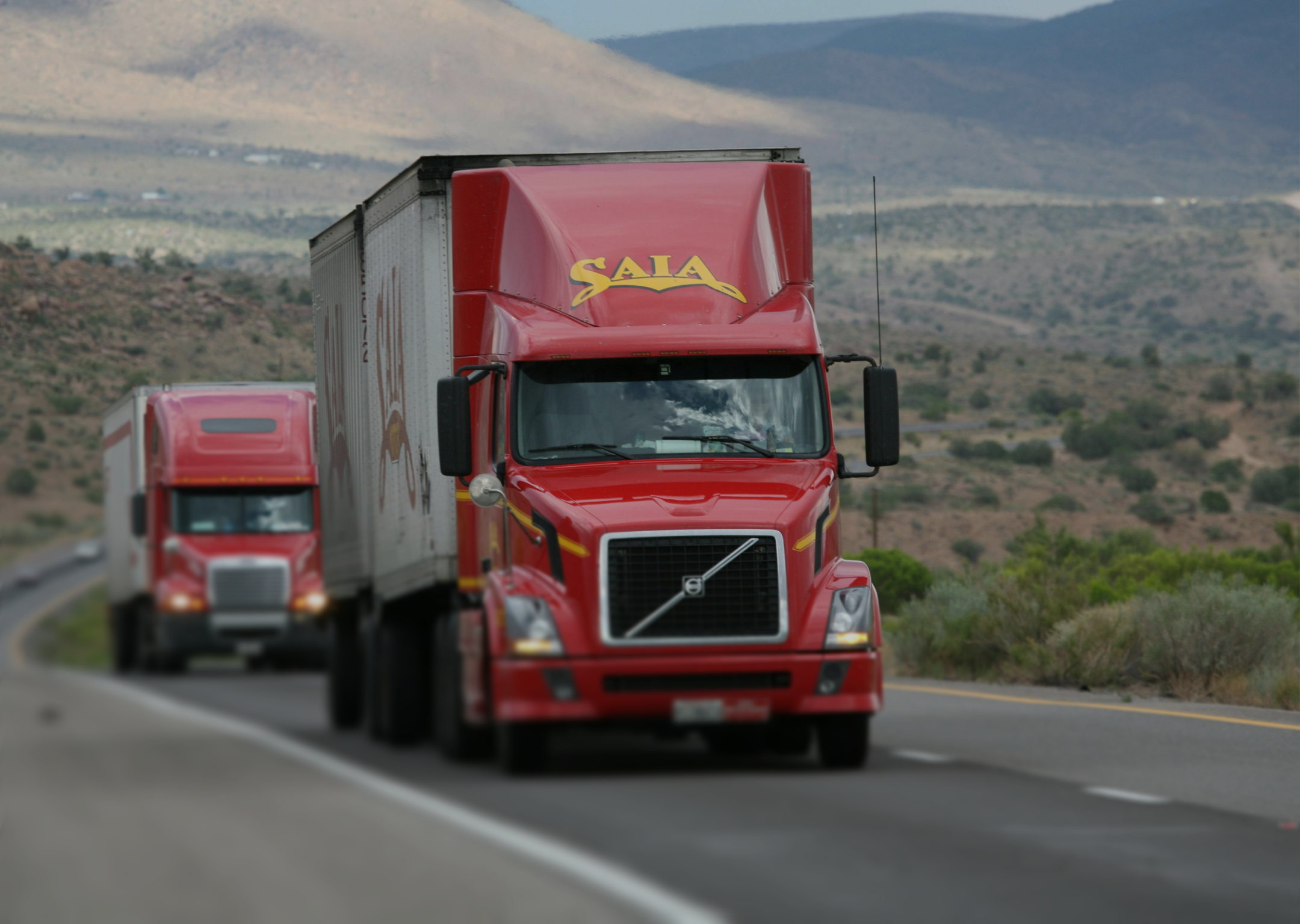 Two red semi-trailer trucks driving on a highway through a mountainous desert landscape.