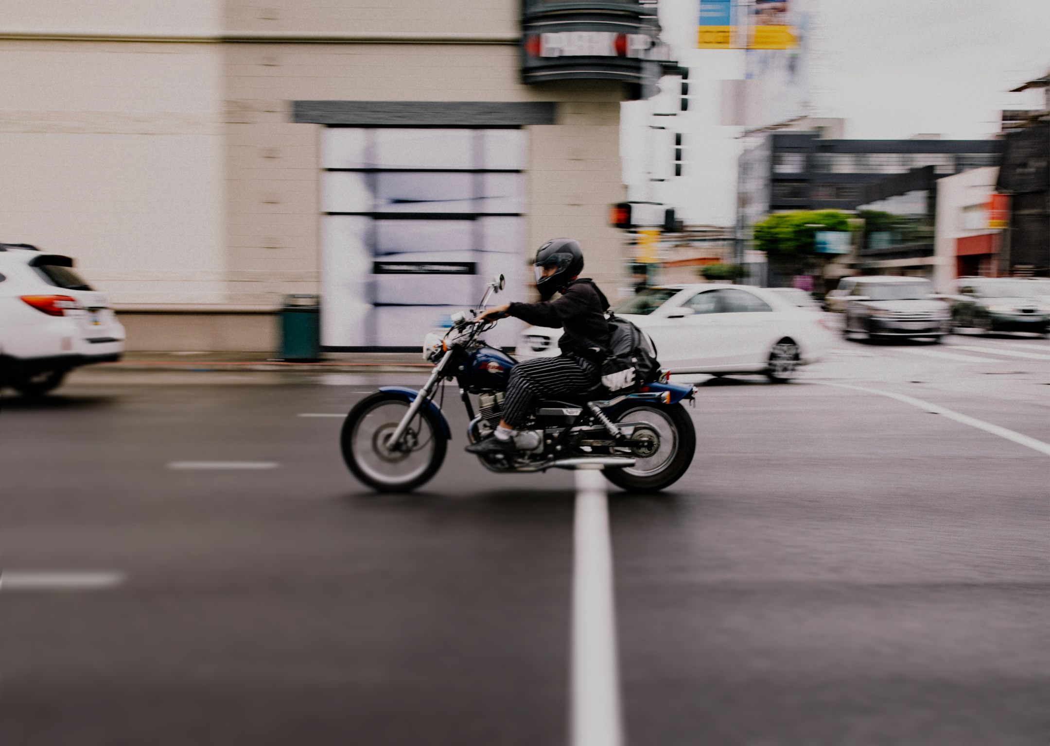 Person wearing a helmet and striped pants riding a blue motorcycle on a city street with blurred cars in the background.