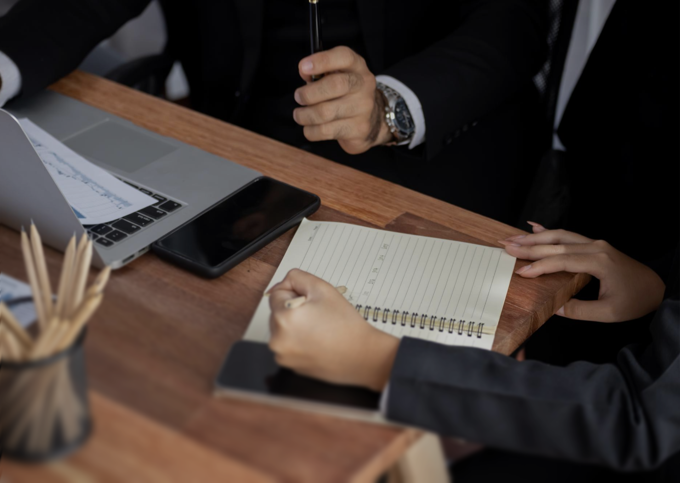 Two people in business attire at a wooden desk with a laptop, smartphone, and a notebook where one person is writing.
