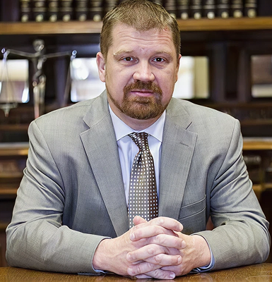 Man with folded hands wearing a light gray suit and polka dot tie, sitting at a wooden table in front of shelves with trophies and books.