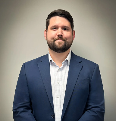 Man with short dark hair and beard wearing a navy blazer and white shirt against a plain light background.