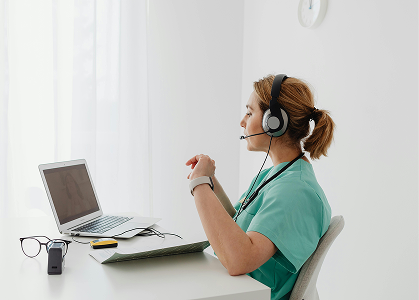 Healthcare professional in scrubs wearing a headset and sitting at a desk with a laptop and medical tablet.