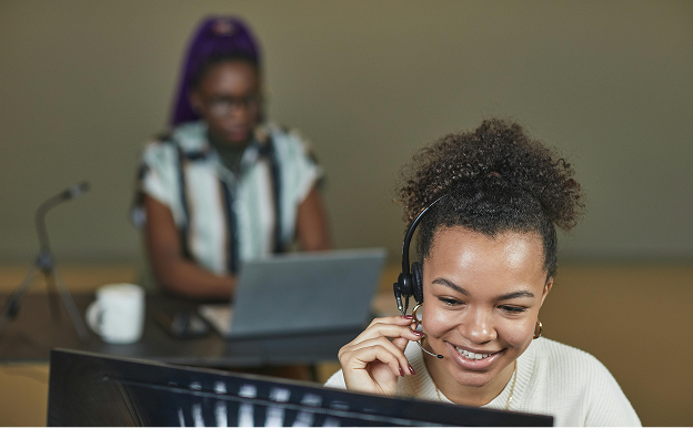Smiling woman wearing a headset working at a computer, with another person using a laptop in the background.