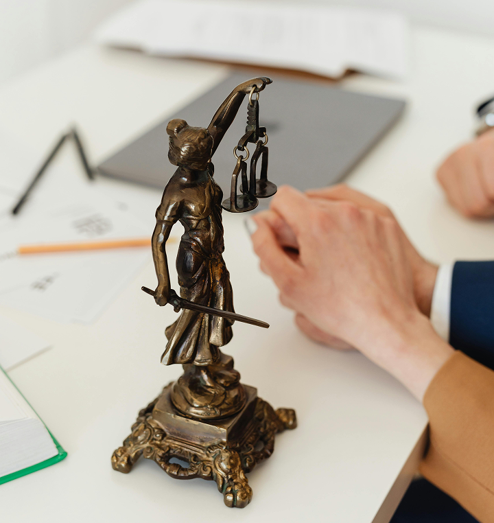 Bronze Lady Justice statue holding scales and sword on a desk with blurred hands and documents in the background.