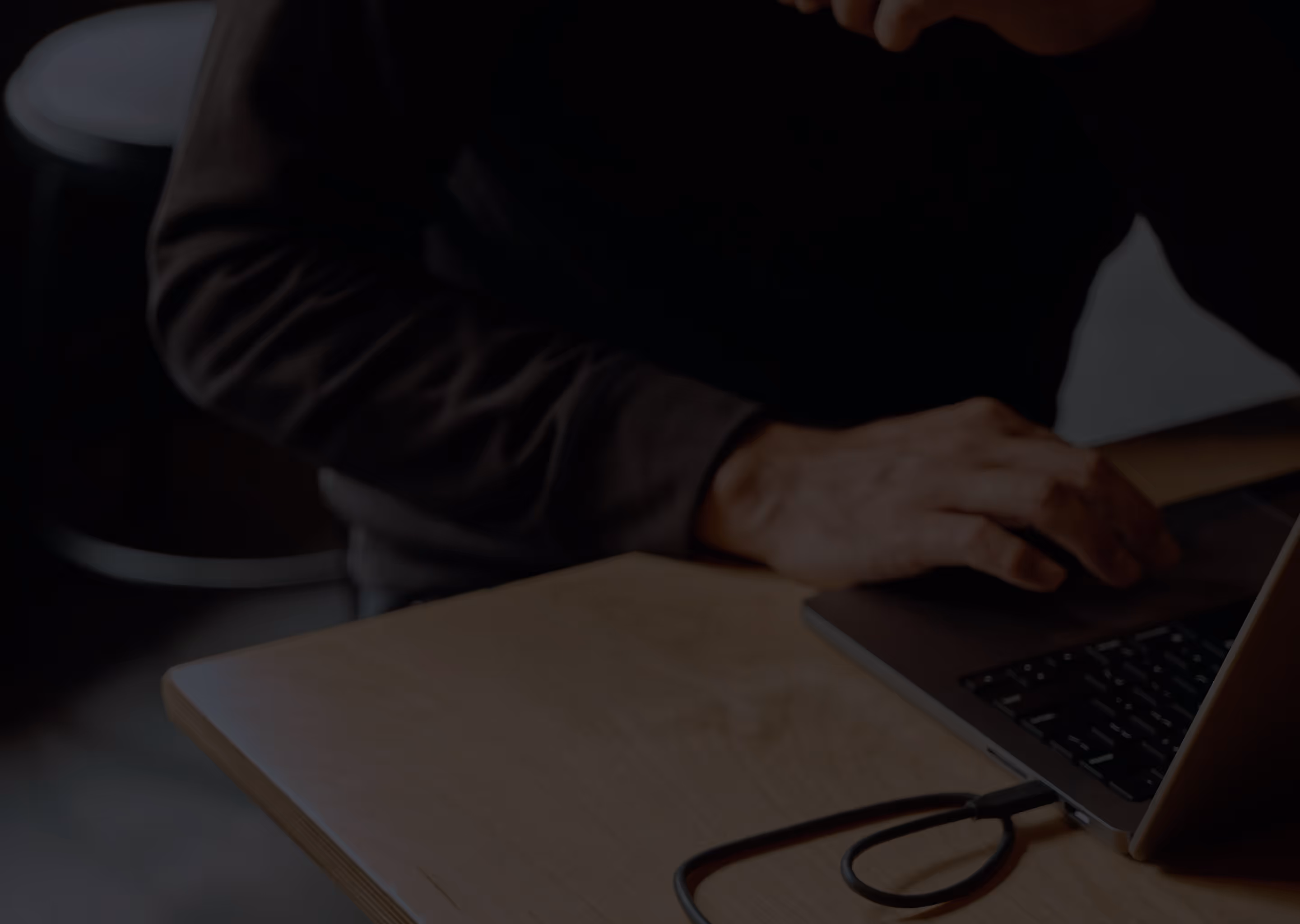 Person typing on a laptop keyboard at a wooden desk with a black charging cable nearby.
