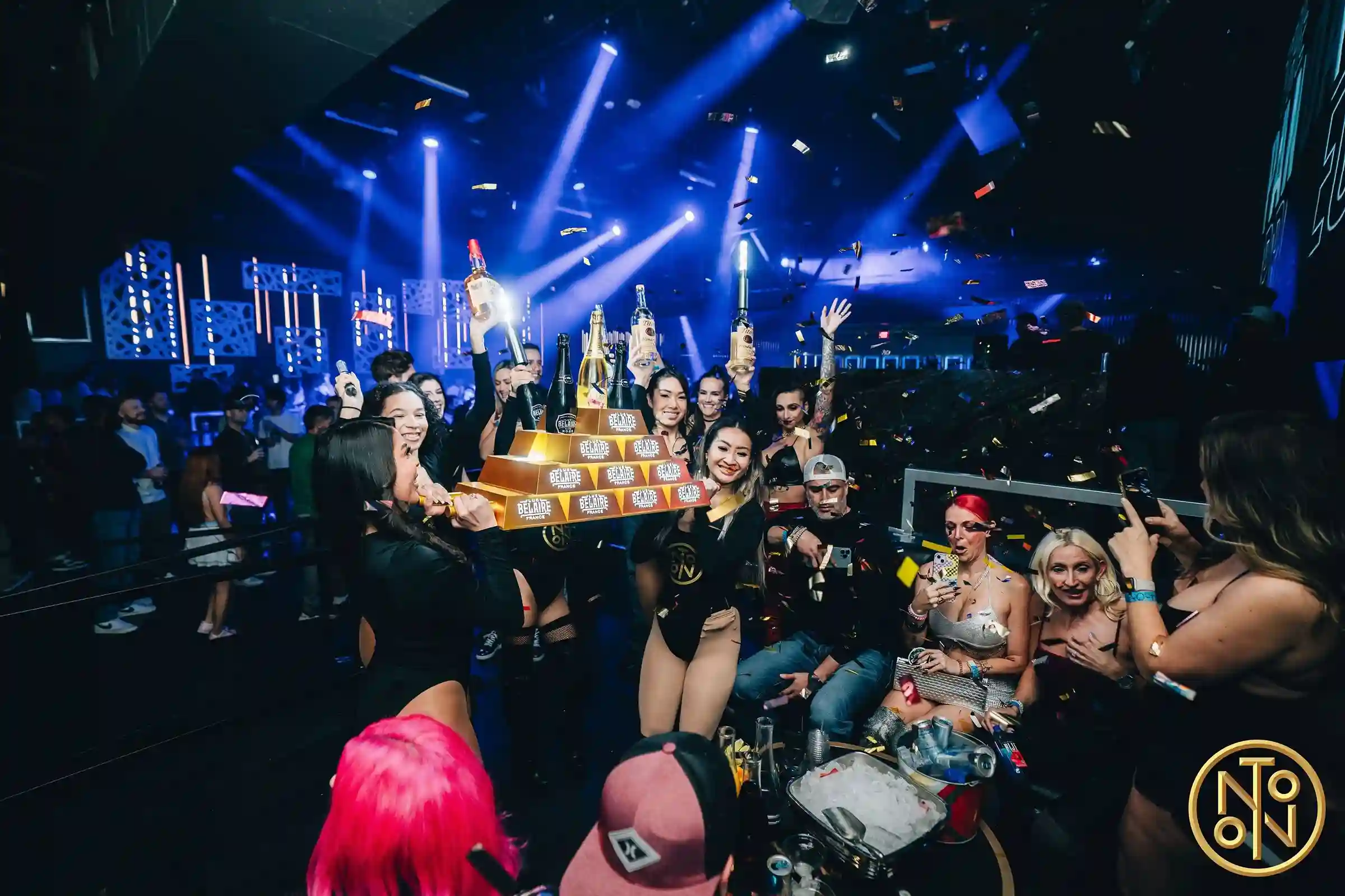 Group of people celebrating in a nightclub with women holding bottles and a golden tray of Belaire French bottles under blue stage lights and confetti.