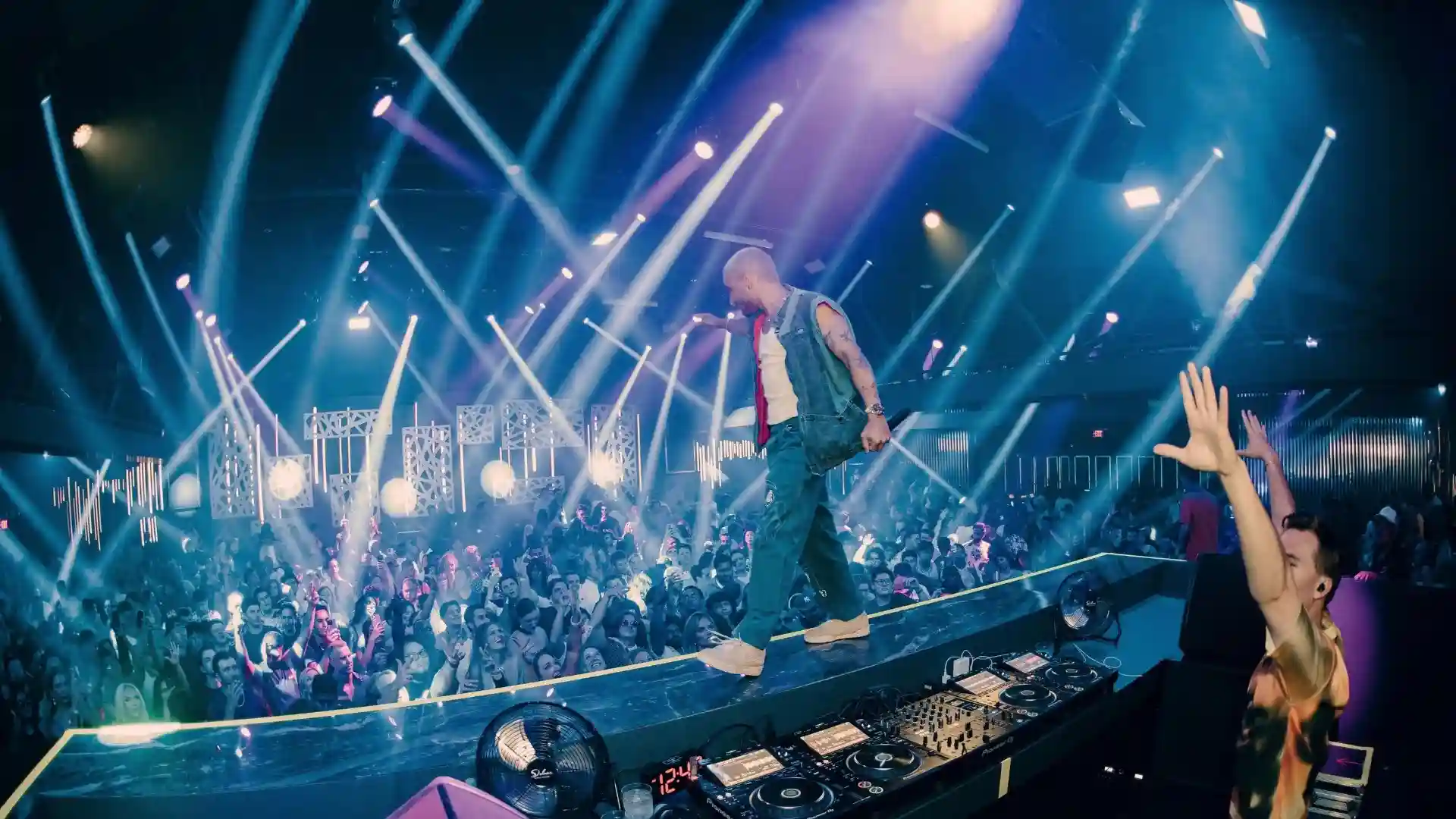 Crowded nightclub with a DJ playing music and a performer walking on the DJ booth under vibrant blue and purple stage lights.
