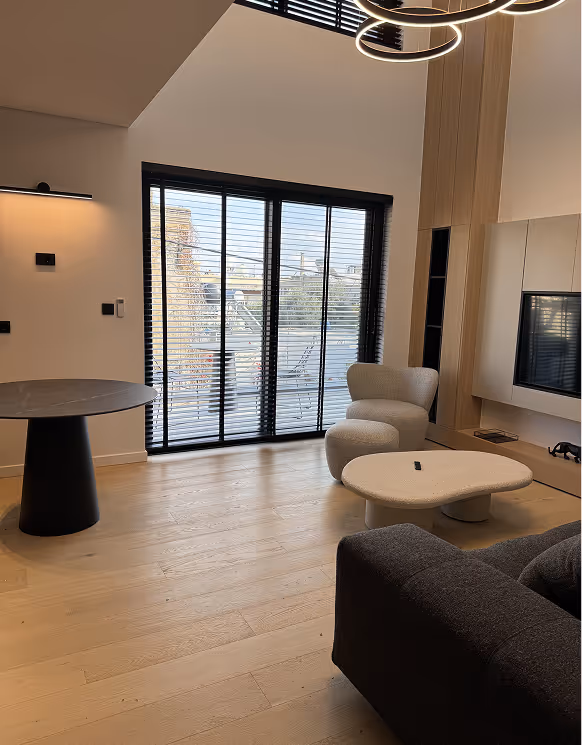 Modern living room with wooden flooring, a round black table, a white curved armchair, a white abstract coffee table, dark sofa, and large window with black blinds overlooking an outdoor patio.