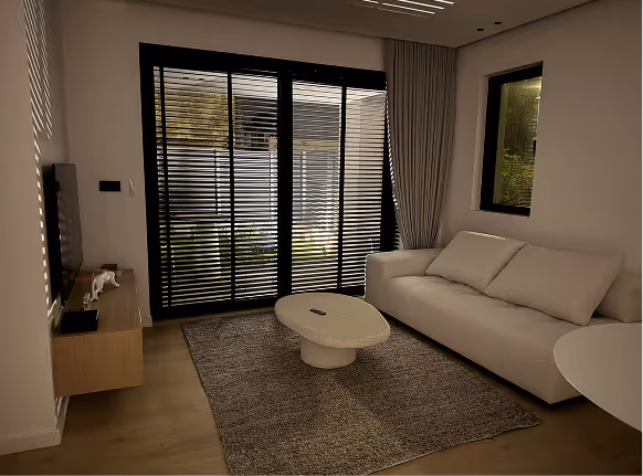Modern living room with a beige sofa, round coffee table on a textured rug, window with curtains, and black horizontal blinds partially open.