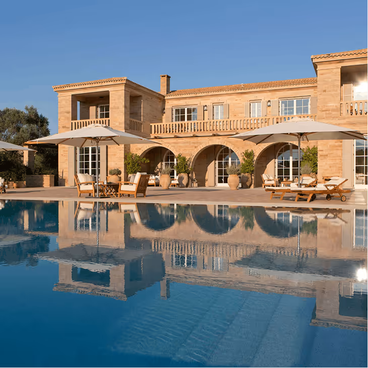 Luxury stone villa with arches, outdoor seating, umbrellas, and a large reflecting pool under clear blue sky.