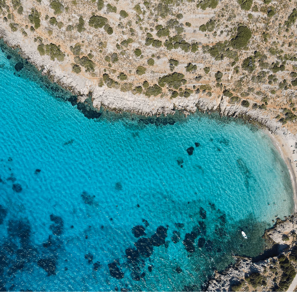 Aerial view of a clear turquoise coastal bay with rocky shoreline and scattered green shrubs.