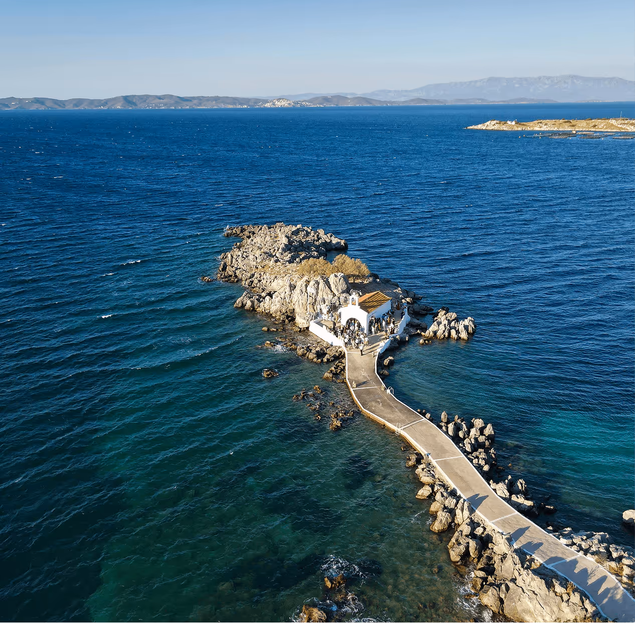 A small white chapel with a tiled roof on a rocky islet connected by a narrow walkway surrounded by blue sea.