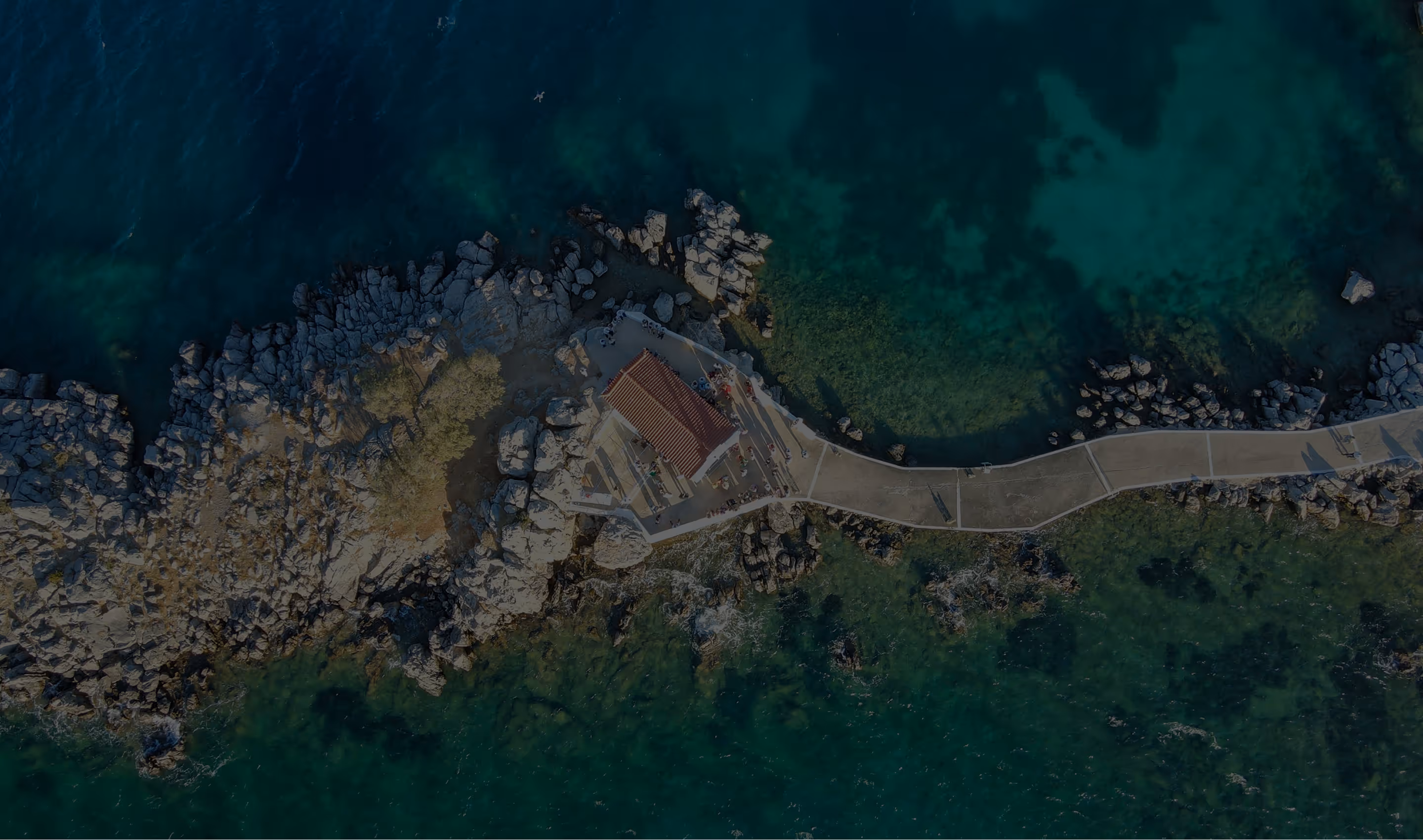 Aerial view of a small building with a red roof on a rocky peninsula surrounded by clear greenish-blue water.