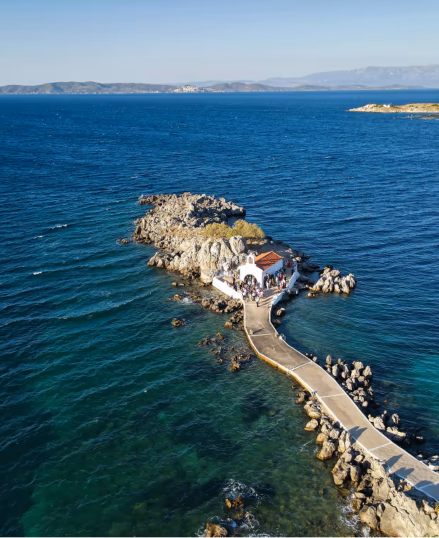 Aerial view of a small white chapel located on a rocky islet connected by a narrow concrete pier, surrounded by blue ocean waters.