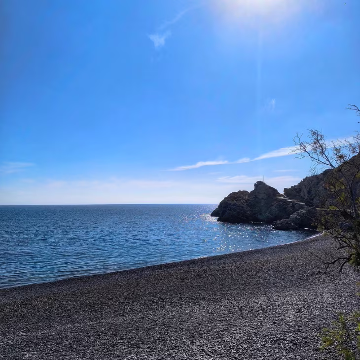 Rocky beach under clear blue sky with calm sea and sunlight reflecting on the water near rocky cliffs.
