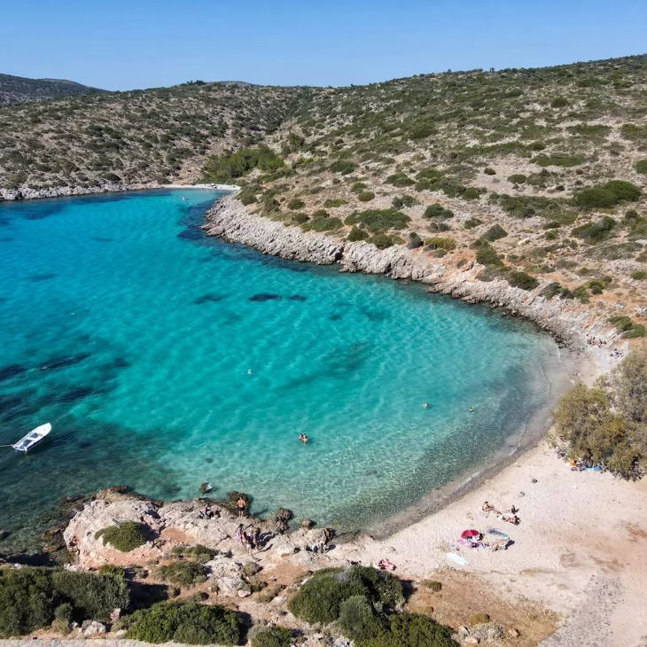 Aerial view of a rocky coastline with turquoise water and a small sandy beach with people sunbathing and swimming.