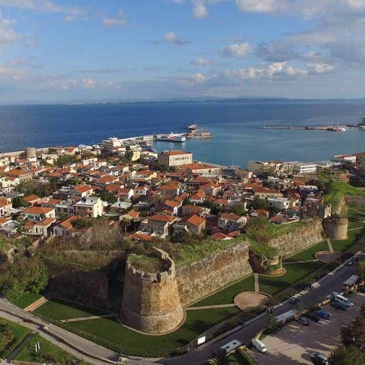 Aerial view of a coastal town with red-roofed buildings, a historic stone fortress wall, and a harbor with ships in calm blue water under a partly cloudy sky.
