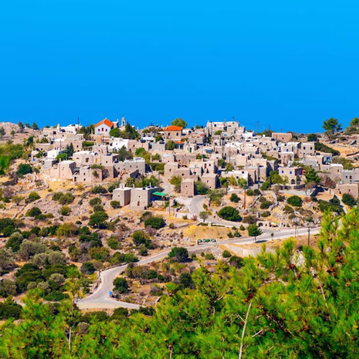 Hilltop village with white stone houses and winding roads surrounded by green trees under a clear blue sky.