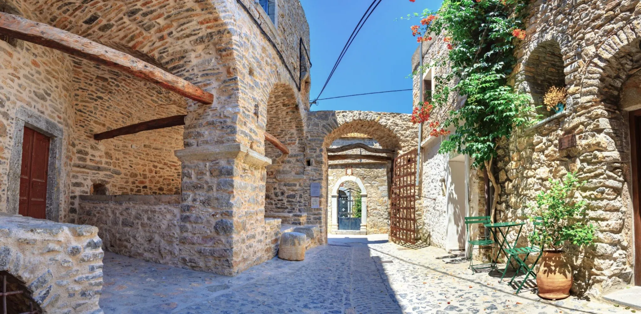 Sunny stone courtyard with arched doorways, wooden beams, green chairs, and potted plants under a clear blue sky.