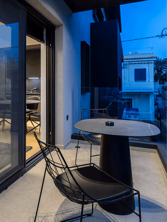 Small outdoor balcony with a round table, two black wireframe chairs, and a view of nearby buildings at dusk.