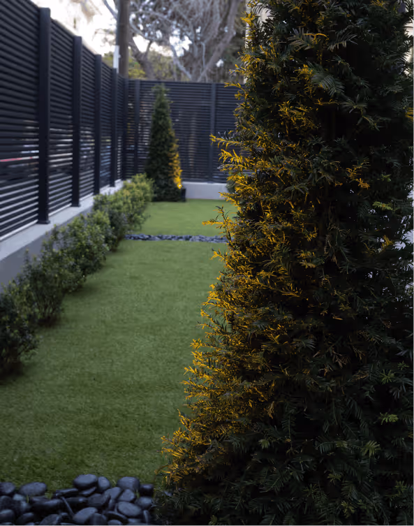 Garden area with artificial green grass, neatly trimmed bushes along a black slatted fence, and tall conical evergreen trees.