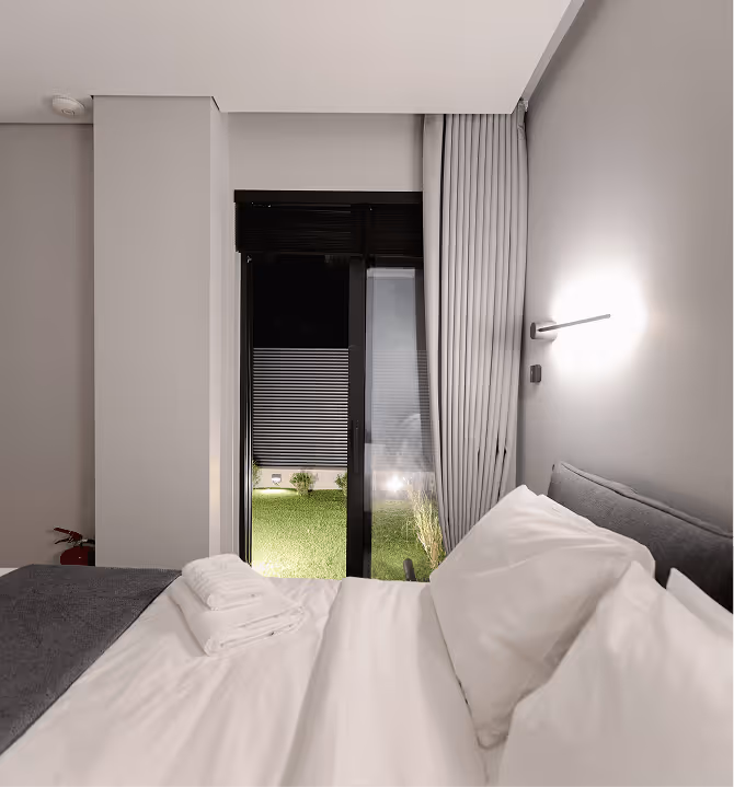 Modern bedroom with neatly made bed, white pillows, folded towels, and sliding glass door opening to a small green lawn.