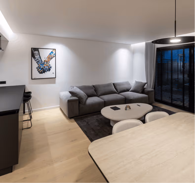 Minimalist living room with a white sofa, white oval coffee table on a gray rug, black window blinds, and light-colored curtains.