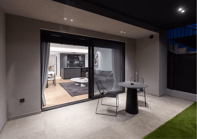Modern patio at night with two black wireframe chairs and a round black table, sliding glass doors open to a living room with a gray sofa and kitchen in the background.