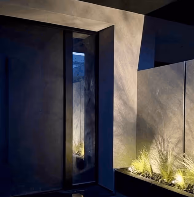 Modern minimalistic corner with tall narrow mirror reflecting lit plants with shadows on concrete walls at night.