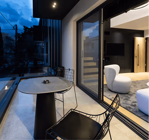 Modern balcony with round black metal table and two black wire-frame chairs overlooking outdoor evening sky, adjacent to living room with white armchairs.