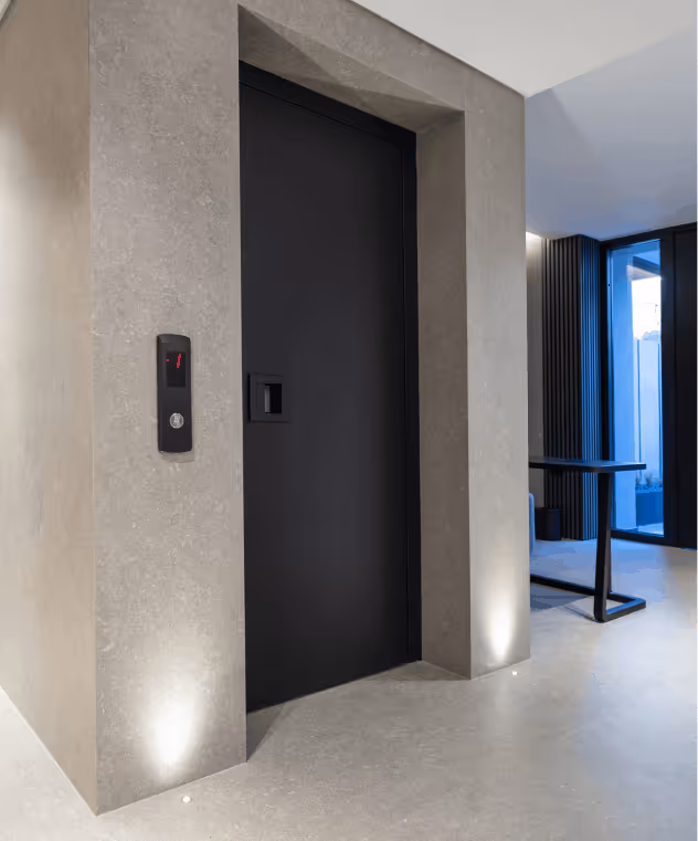Modern interior with a closed black elevator door set in a light grey textured wall with recessed floor lighting and a sleek elevator control panel.