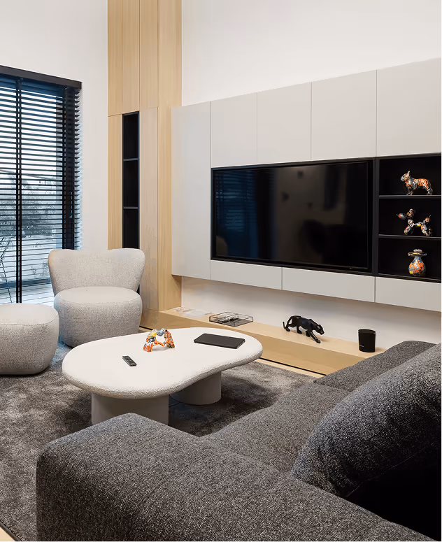 Modern living room with gray sofa, rounded white coffee table, gray armchair, and wall-mounted TV with decorative shelves.