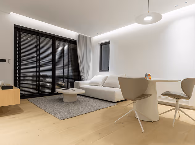 Minimalist living room with a white sofa, gray rug, round coffee table, dining table with two chairs, and black window blinds.
