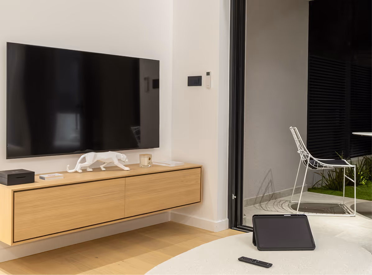 Modern living room corner with a wall-mounted flat-screen TV above a wooden floating cabinet, a white ceramic panther sculpture, and a tablet and remote on a light surface next to a sliding glass door leading to an outdoor area with a white metal chair.