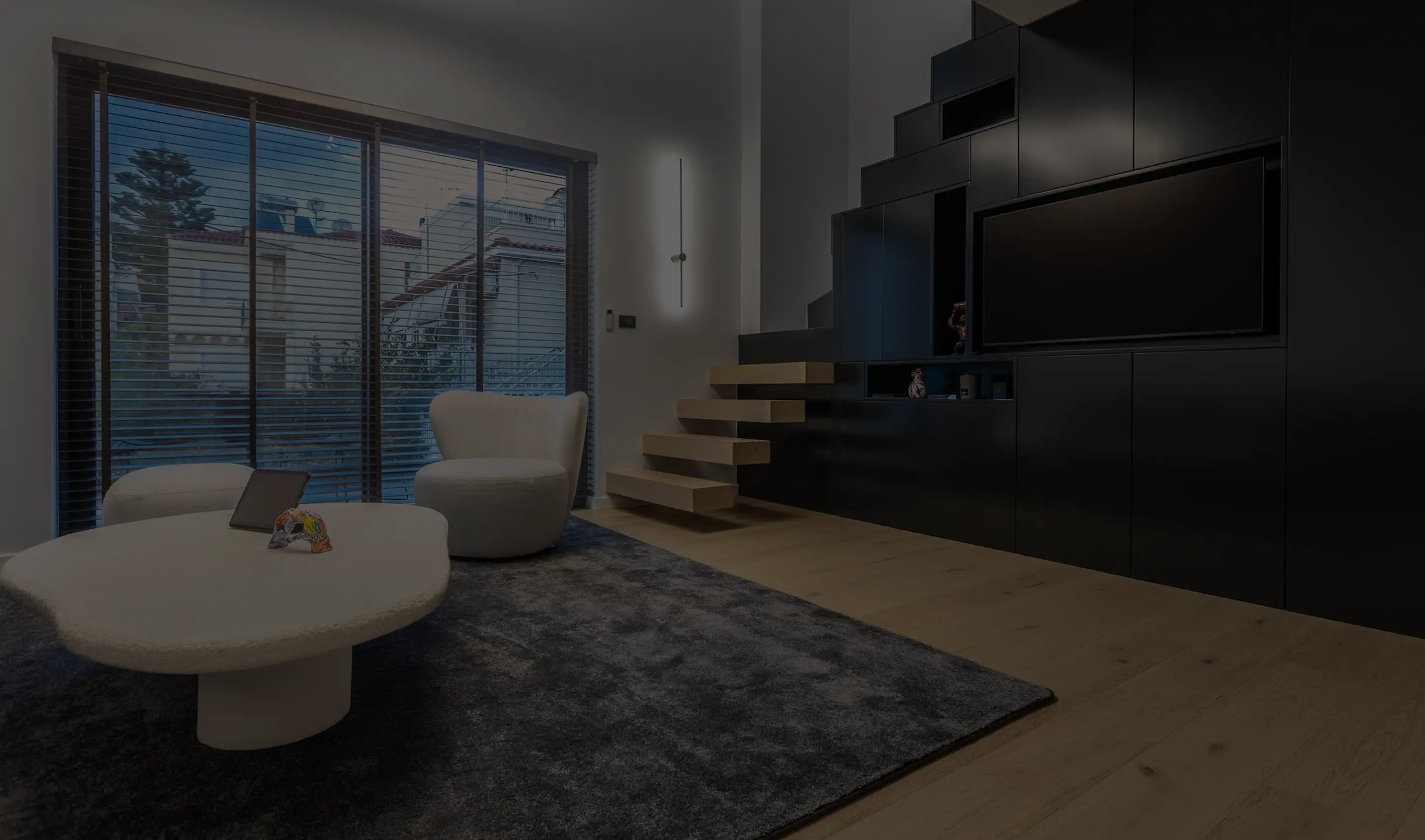 Modern living room with a white lounge chair, matching ottoman, irregular white coffee table, floating wooden stairs, black entertainment wall with TV, and large window with horizontal blinds.