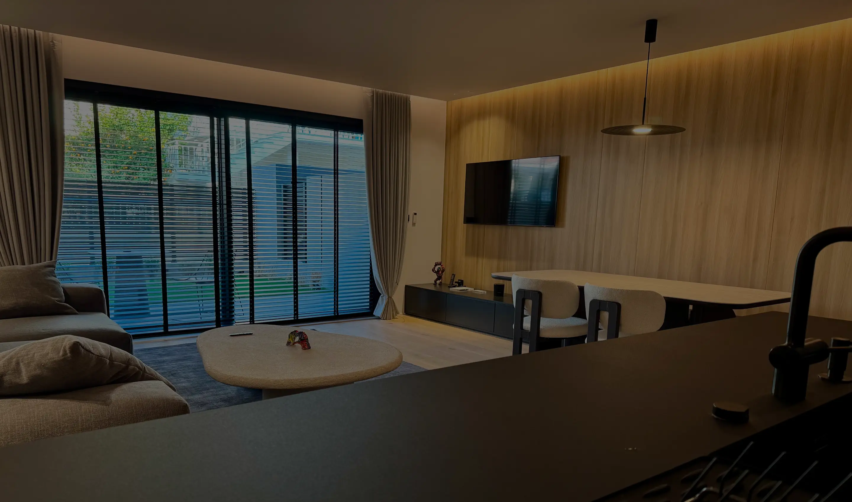 Modern living room with large window blinds, beige sofa, triangular coffee table, wall-mounted TV, dining table with chairs, and pendant light.