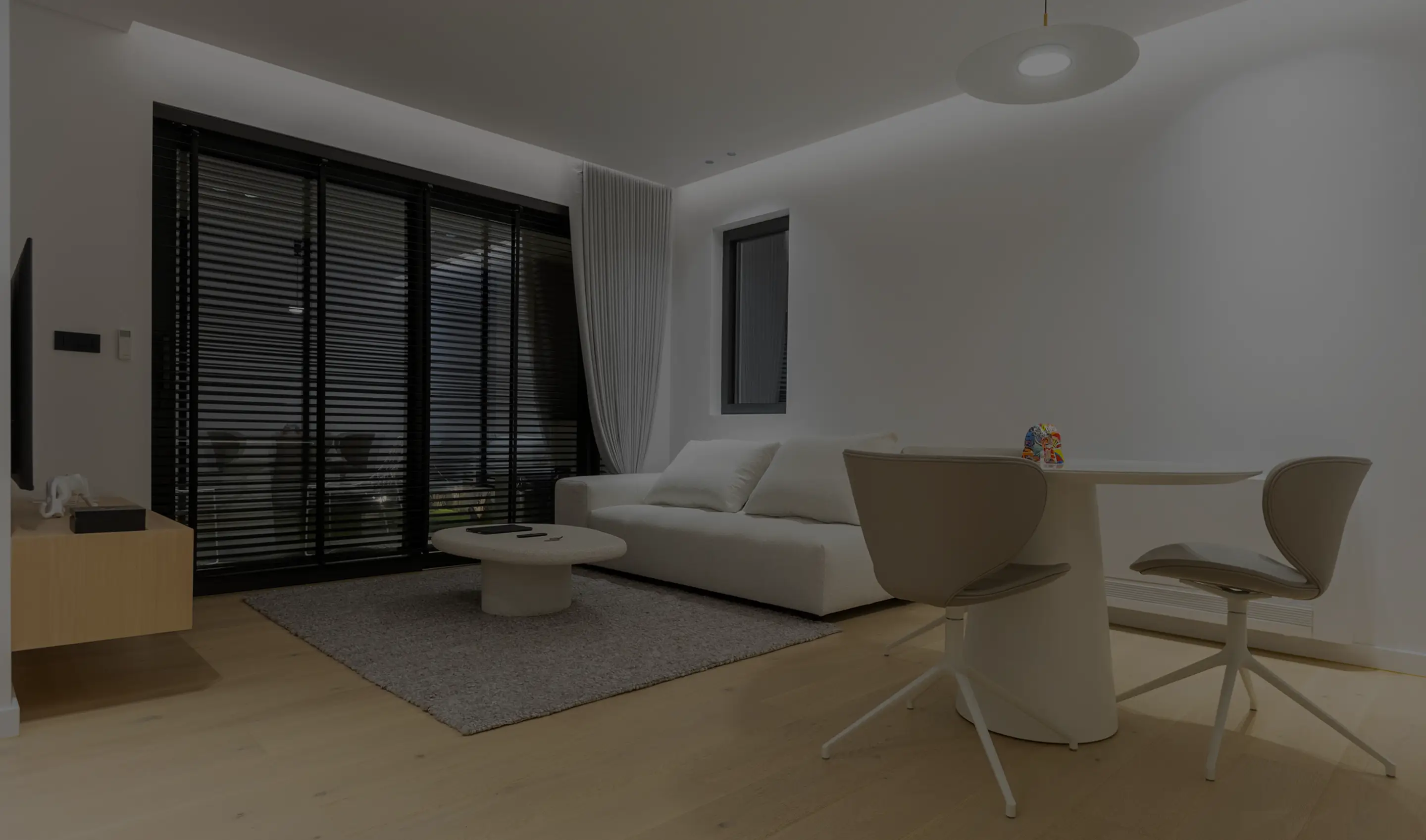 Modern living room with a white sofa, round coffee table on a rug, flat-screen TV on a wooden stand, and a small dining table with two chairs.